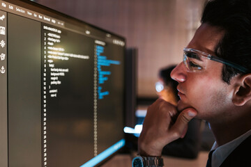 Focused man in glasses analyzes code on a computer monitor in a modern office, representing software development, programming concentration, and tech-driven problem-solving in a digital workspace.