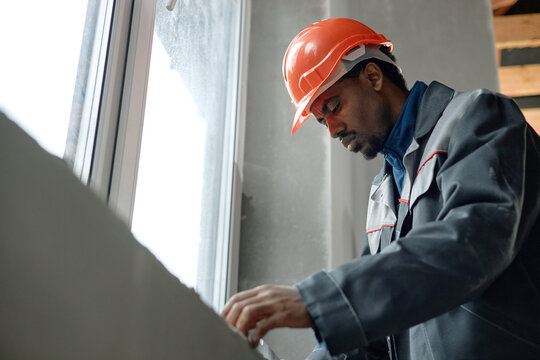 Black young adult man wearing construction helmet working near window, focusing on measuring or adjusting surface, demonstrating concentration and manual skill in industrial setting