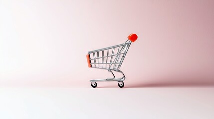 A miniature metal shopping cart with orange handles sits on a pink surface, isolated against a soft pink background.