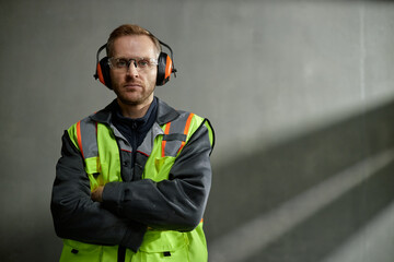 Portrait of Caucasian middle aged man wearing safety vest and protective earmuffs standing with arms crossed, looking into camera in industrial setting with concrete wall background