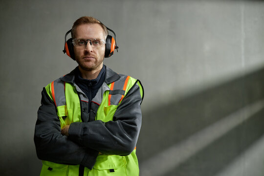 Portrait of Caucasian middle aged man wearing safety vest and protective earmuffs standing with arms crossed, looking into camera in industrial setting with concrete wall background