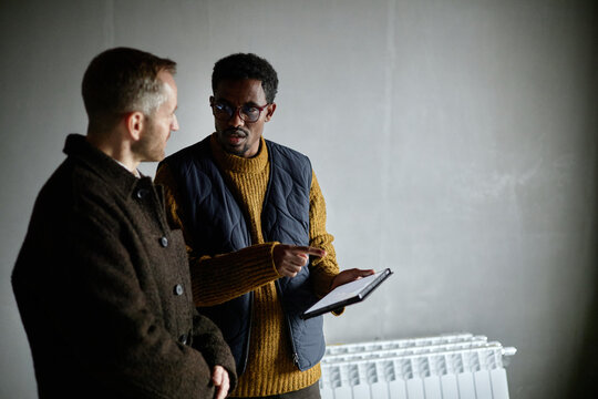 Middle aged Caucasian man and middle aged Black man standing together discussing information on digital tablet, Black man gesturing while explaining details to colleague