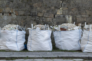 Worki big bag wypełnione gruzem budowlanym stojące przy murze miejskiej ulicy.