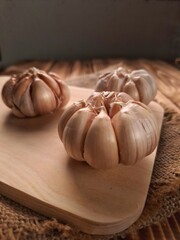 Three garlics bulb on top a woodeboard and a rug. Three garlic bulbs rest on a light-colored wooden board, placed on a textured burlap fabric.
