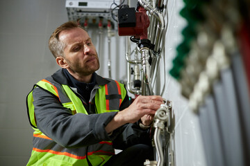 Caucasian middle aged man inspecting industrial pipes and adjusting valves in technical room, wearing safety vest and focusing on maintenance work, modern equipment visible in background