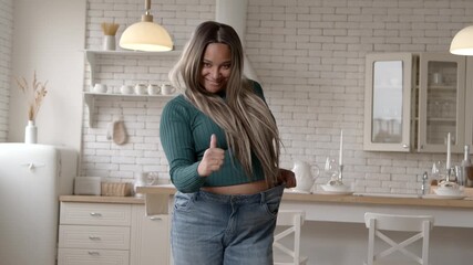 Charming body positive plus size African American woman in oversized jeans celebrating results of successful slimming program, showing thumbs up, expressing excitement and joy at home.
