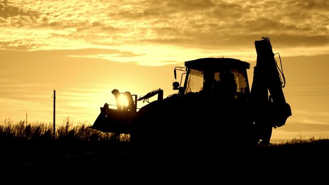 Worker working on bulldozer, backlit silhouette of powerful building machinery. Beautiful sunset or sunrise in countryside, professional dozer driver or famer, earthworks and engineering works