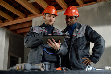 Caucasian young adult man and Black young adult man wearing protective helmets collaborating and reviewing construction plans on digital tablet at building site