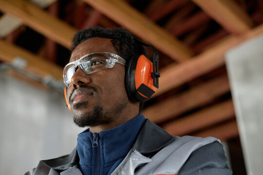 Portrait of young adult Black man wearing protective earmuffs and safety glasses working in industrial setting, looking focused and attentive, visible wooden beams in background