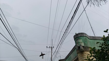 A view of tangled overhead power lines stretching across a cloudy sky near a residential building. This photo is ideal for illustrating urban infrastructure, electrical networks, public utilities, and