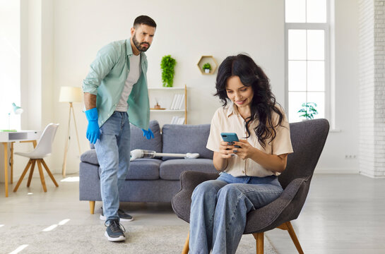 Couple at home doing cleaning chores together. In a sunny living room, a man vacuums near the sofa while a woman sits in a chair using a smartphone. Modern family roles and responsibilities.