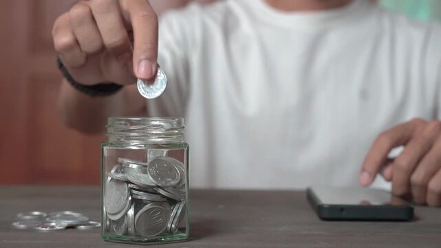 A young Asian man in a white shirt sits with a piggy bank, coins, a calculator, and a cell phone. He is focused on financial stability, saving for emergencies, education, and future goals.