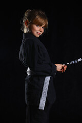 Confident young girl in black martial arts gi with white belt holding a training strap, posing proudly for kids self-defense, BJJ and martial arts lifestyle concepts.
