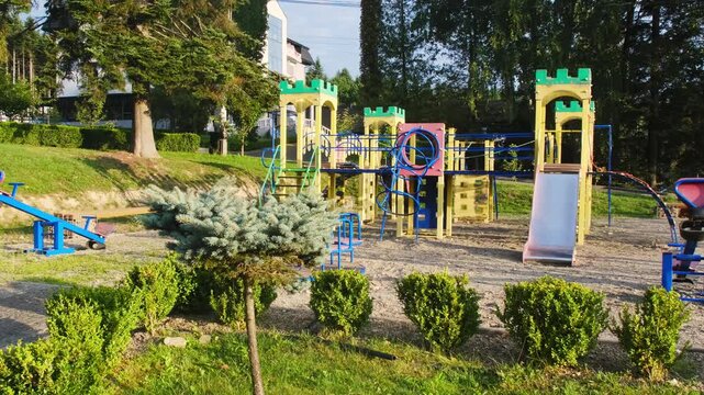 Colorful playground shows slides ladders bridges and towers under pine trees. Fresh gravel covers ground and shrubs line border near quiet path.