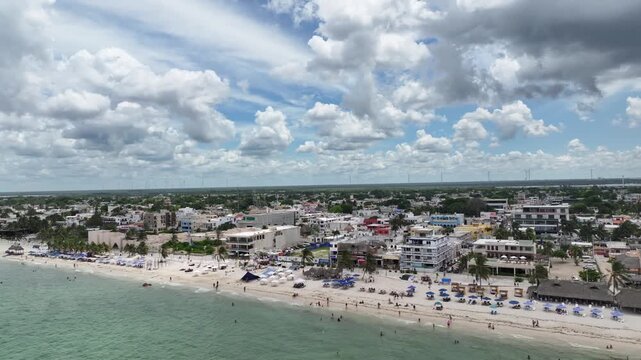 A lateral drone flight over Puerto Progreso, showing the beachfront city with crowded sandy shores, turquoise water, and dramatic towering clouds on a bright sunny day.