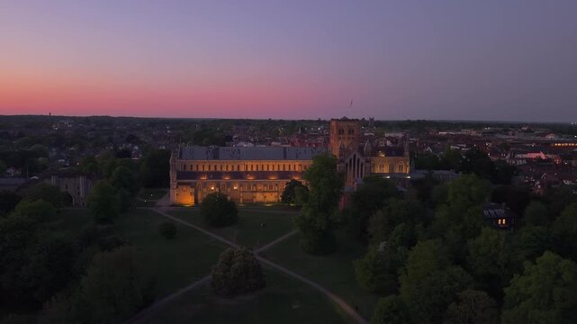 Aerial drone shot captures the illuminated St Albans Cathedral at twilight, gradually revealing the glowing city lights behind and the serene evening atmosphere