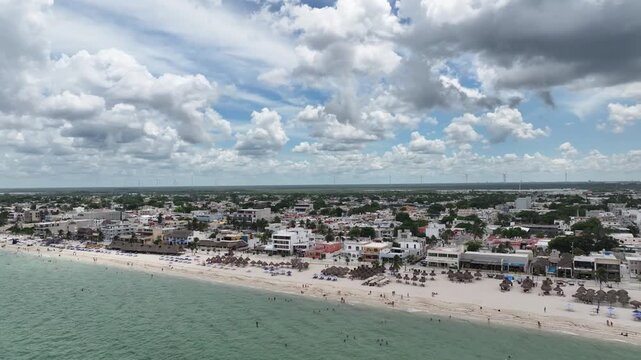 A lateral drone flight over Puerto Progreso, showing the beachfront city with crowded sandy shores, turquoise water, and dramatic towering clouds on a bright sunny day.