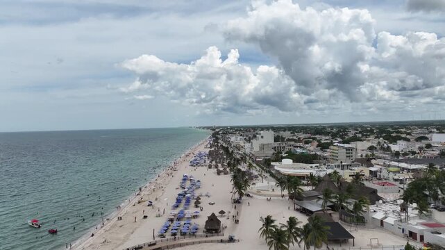 Drone flying forward along the crowded beach of Progreso, Yucat&aacute;n, showing turquoise water, umbrellas, and the city under dramatic clouds.