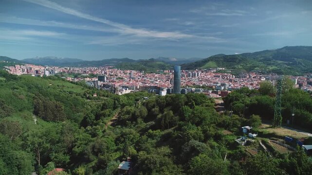 Aerial drone tracking shot over a green hillside looking toward Bilbao city skyline with the Iberdrola Tower and surrounding mountains on a clear sunny day.
