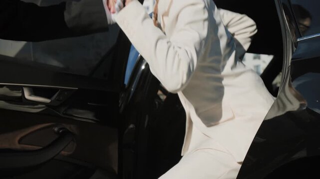 Professional male personal driver in white gloves helping businesswoman leaving car while arriving at business venue in city street