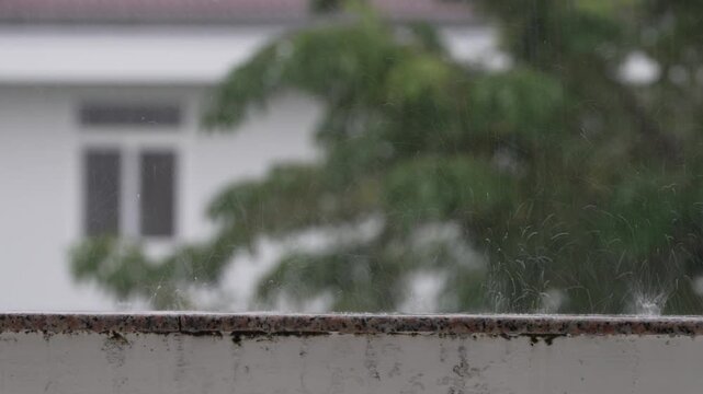 Raindrops falling on a stone balcony railing, blurry garden and house in the background