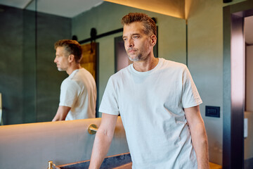 Naklejka premium Thoughtful man looking away in a contemporary bathroom, reflecting in a mirror. Concepts of contemplation, wellness and morning routine