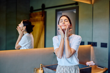 Woman standing in bathroom, gently massaging moisturizer into her face with her eyes closed, practicing self-care