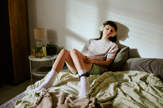 Young adult biracial woman sitting on bed wearing electronic ankle monitor and headphones, relaxing at home with eyes closed, sunlight casting shadows on wall, hands resting on legs