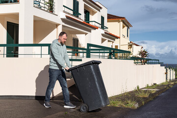 Man pushing black trash bin along sidewalk near residential buildings with greenery