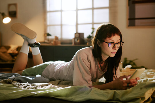 Young adult Asian woman lying on bed using smartphone at home wearing electronic ankle monitor, staying indoors under house arrest concept modern monitoring technology visible