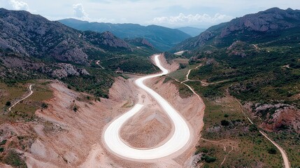 An aerial view shows a white, winding road snaking through a dramatic mountain valley with steep, rocky slopes and patches of green vegetation.
