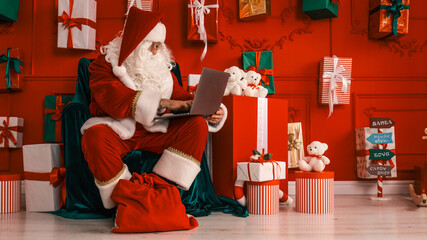 Santa Claus working on a laptop surrounded by Christmas presents and decorations