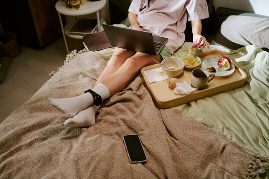 Unrecognizable woman sitting on bed using laptop with ankle monitor visible on leg, eating snacks from tray, staying at home with electronic monitoring device