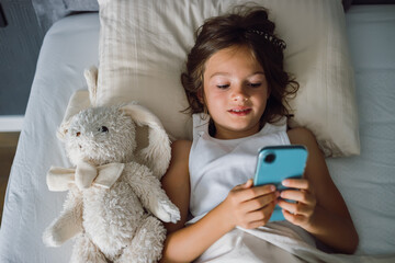 Young girl lying in bed with her plush toy, using a smartphone, illustrating modern childhood, screen time, and children's engagement with technology