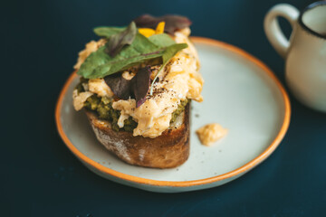 Close-up of a sourdough toast topped with creamy scrambled eggs, fresh avocado, and greens, served on a plate against a dark table background