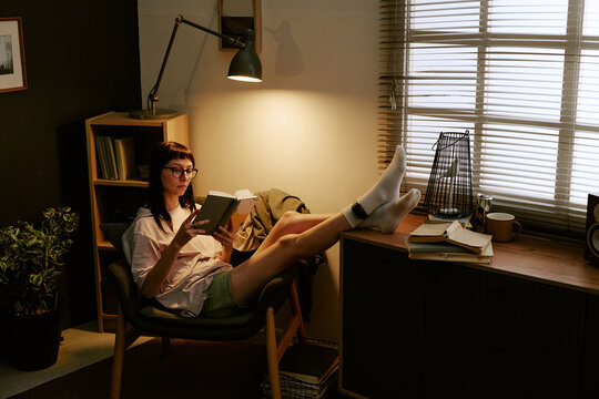 Young adult biracial woman sitting in chair reading book at home with electronic ankle monitor visible on leg, relaxing in living room near window with natural light coming through blinds