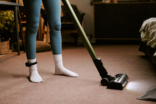 Unrecognizable woman vacuuming carpet at home with electronic ankle monitor visible on left leg, demonstrating daily life under house arrest or electronic monitoring