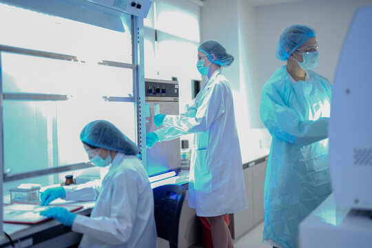 Three female scientists in protective gear work in a modern laboratory. They are operating machines and working in biosafety cabinets during a busy day of research and testing. - Powered by Adobe