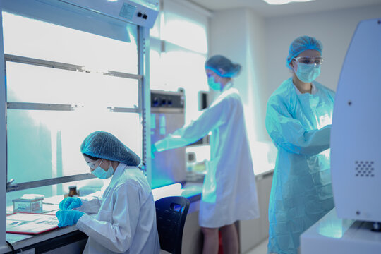Three female scientists in protective gear work in a modern laboratory. They are operating machines and working in biosafety cabinets during a busy day of research and testing.
