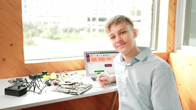 Smart adult repair main board while turn around to look at camera with confident on table with laptop with engineering code displayed screen and electronic equipment at STEM classroom. Edification