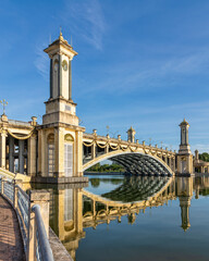 Grand Seri Gemilang Bridge with ornate towers and clock faces, reflected in calm water under a blue sky in Putrajaya, Malaysia