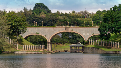 Stone arch bridge over Putrajaya Lake, featuring a charming gazebo, lush green hills, and decorative lampposts. Located in Putrajaya, Kuala Lumpur, Malaysia.