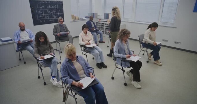Adult Students Sit Individual Desks in Bright, Minimalist Classroom, Writing on Paper Assignments. Female Teacher Walks Between Rows, Answering Questions and Guiding Learners Through Lesson Material.