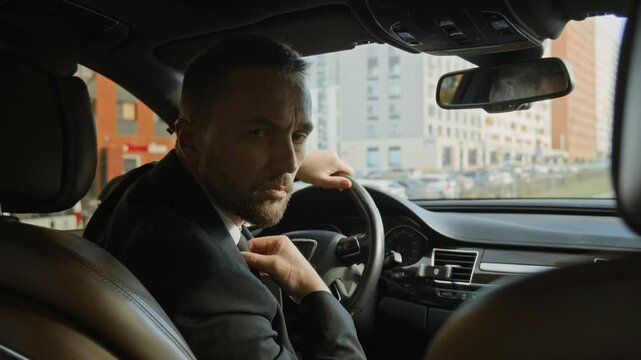 Portrait of middle aged male personal driver in suit sitting in front seat of executive class car handling parking vehicle at business venue, offering premium service to high profile clients