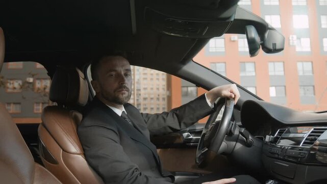 Portrait of middle aged male personal driver in suit adjusting rear view mirror sitting in front seat of luxurious car at parking lot, providing high standard service for business clients