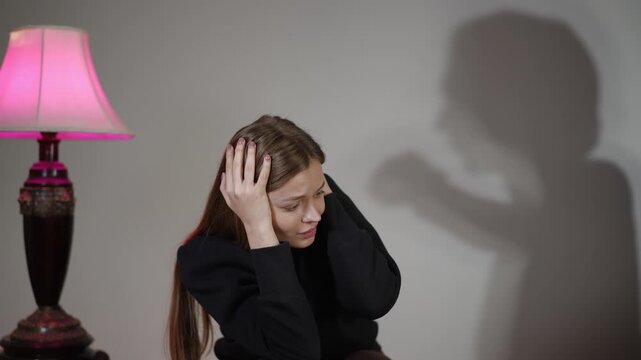 Caucasian woman flinching beside pink lamp as looming male-shaped shadow reaches toward her living room setting, hands covering head, anxious expression,