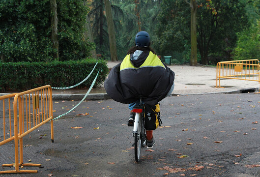 Montando en bicicleta con abrigo poncho, capucha y chubasquero impermeable bajo la lluvia entre &aacute;rboles y vallas