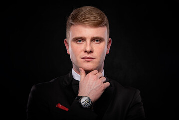 A handsome young Caucasian man in a black tuxedo and bow tie, wearing a wristwatch, holds his chin while looking directly at the camera against a dramatic black background.