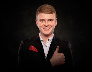 Studio portrait of a smiling young redheaded man wearing a formal black suit and giving a thumbs up gesture of approval against a dark background.