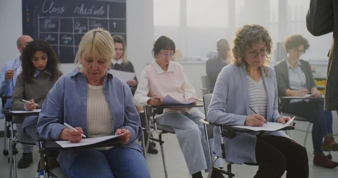 Focused Group Adult Students Writing During Lesson in Classroom. Students Complete Written Exercises Modern Individual Desks. Lifelong Learning, Inclusive Education, Personal Development. Dolly Shot. - Powered by Adobe